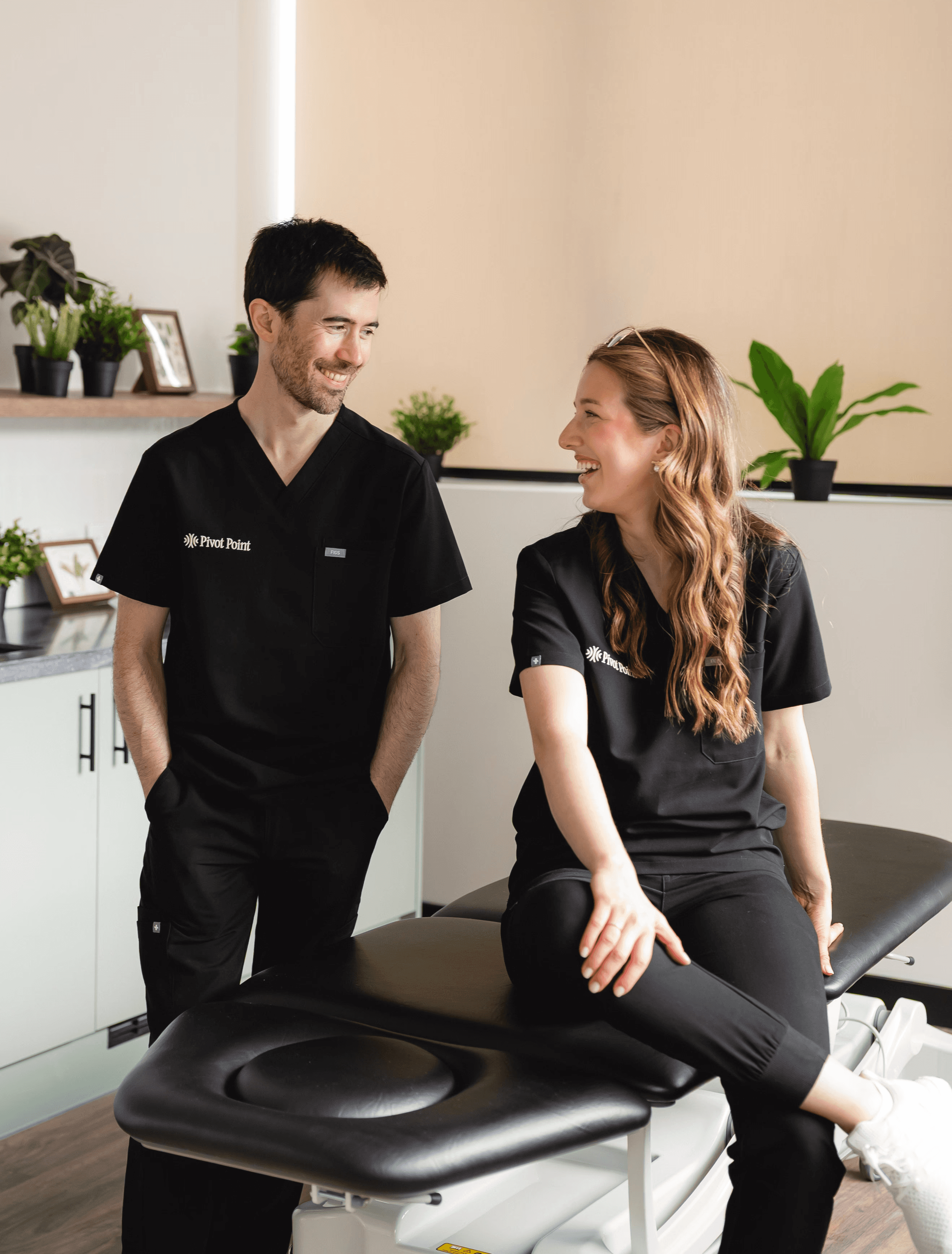 Two physiotherapists in uniform in a treatment room, facing each other and smiling