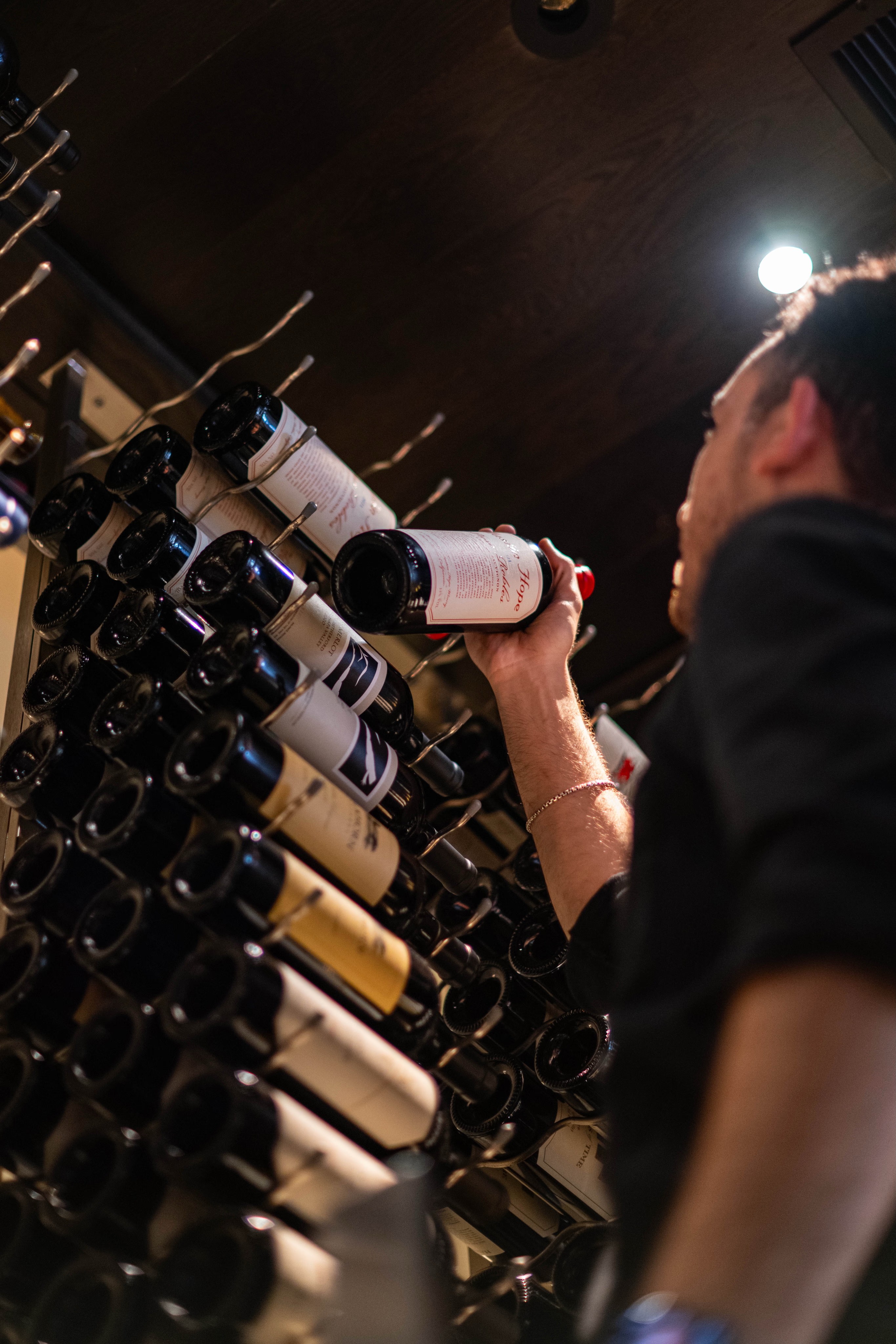 A person holding a wine bottle while examining a rack filled with wine bottles in a dimly lit space.