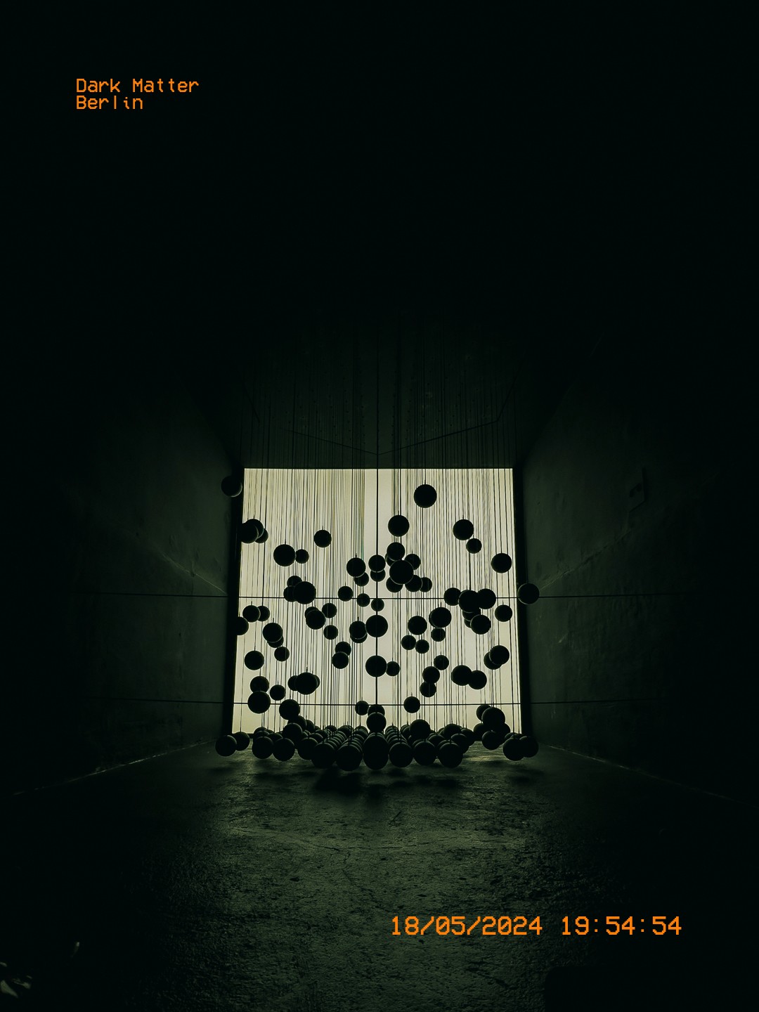 Œuvre cinétique suspendue à Dark Matter Berlin, éclairée par un fond lumineux.