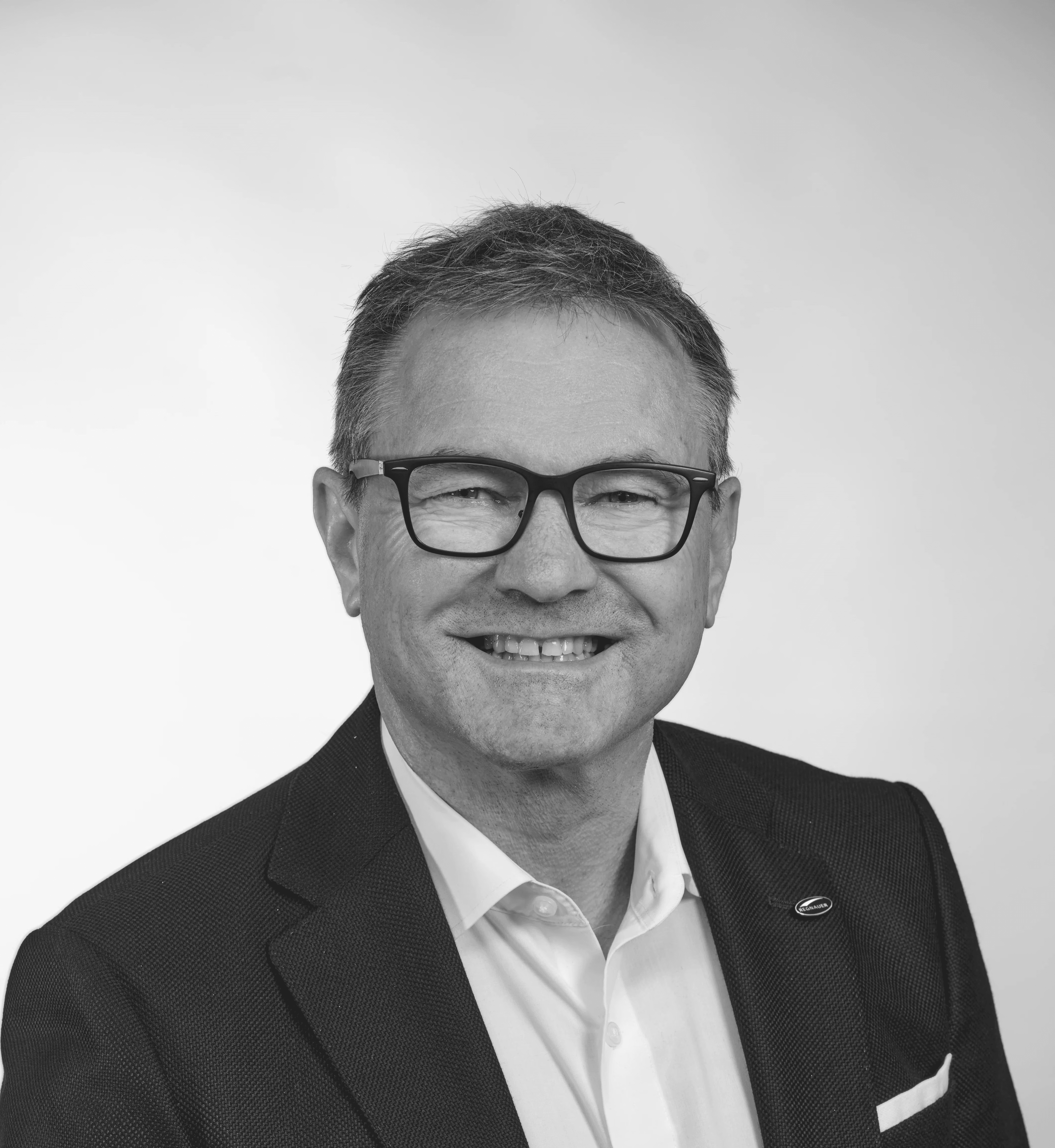 Porträt eines professionell gekleideten Mannes mittleren Alters im Brustbereich, der eine schwarze Brille trägt und in die Kamera lächelt. Er hat graues Haar, eine gepflegte Gesichtsbehaarung und trägt einen dunklen Anzug mit einem weißen Hemd. Am linken Revers des Anzugs ist eine kleine Anstecknadel mit der Aufschrift "REGNAUER" zu sehen.
