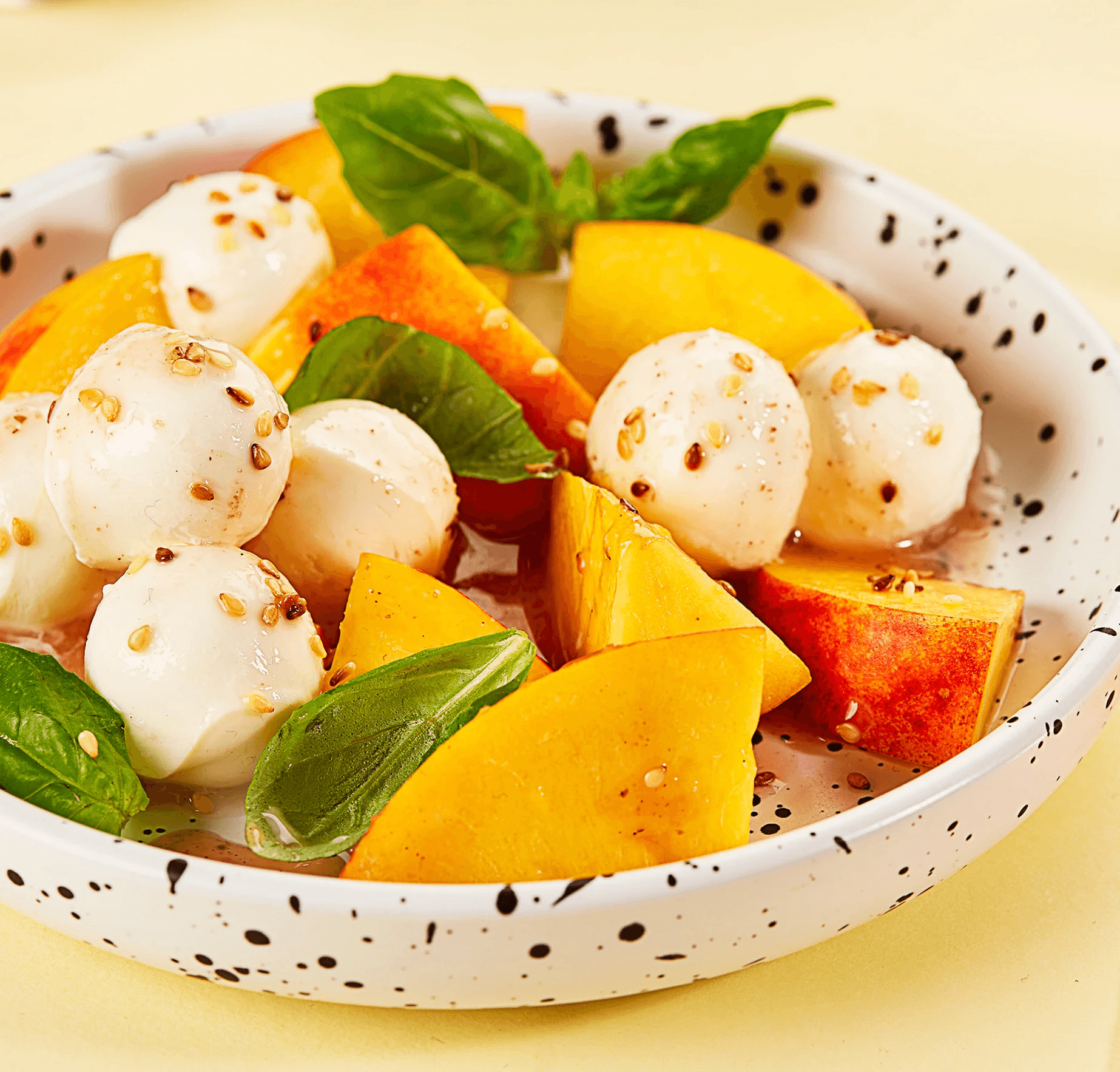 A fresh and vibrant caprese salad features juicy peach slices, creamy mozzarella balls, fragrant basil leaves, and a sprinkle of sesame seeds, all artfully arranged on a speckled white plate.