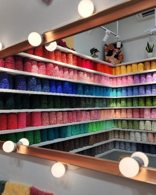 A vibrant wall display of neatly arranged yarn spools in various colors, including pink, blue, green, and orange, reflected in a mirror with round lights.