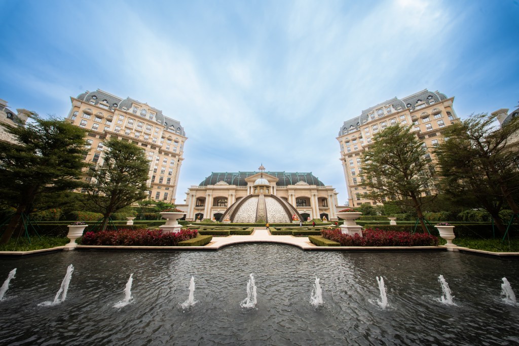 A luxurious hotel entrance with a large fountain in the foreground, surrounded by landscaped gardens and elegant buildings under a clear blue sky.