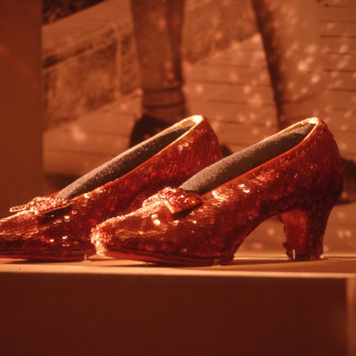 รองเท้าส้นสูงสีแดงประดับเลื่อมพร้อมโบว์ จัดแสดงบนพื้นหลังเบลอซึ่งมีภาพขาวดำ
