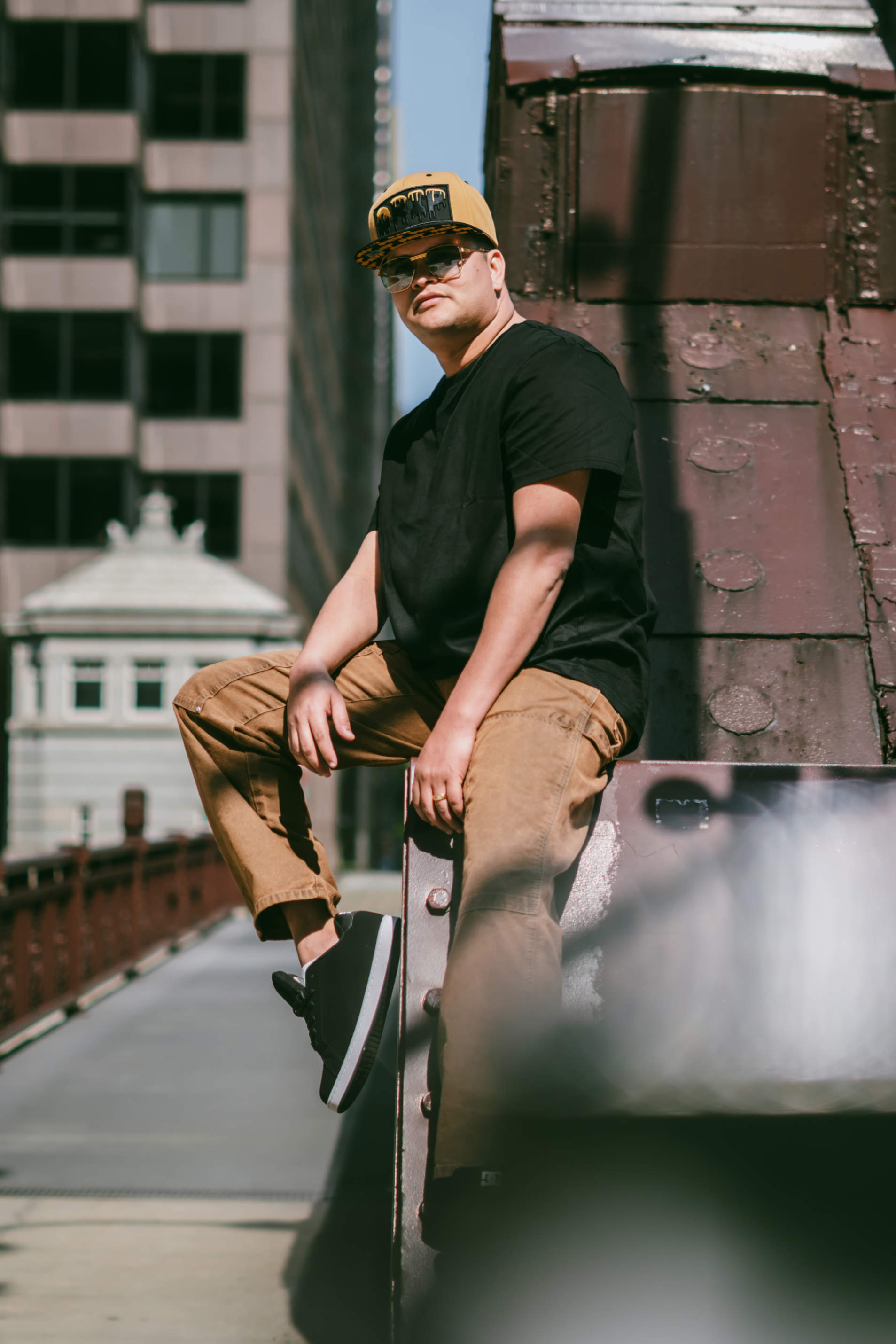 Dani Sierra in casual attire sits on a ledge, with urban buildings in the background on a bright day.