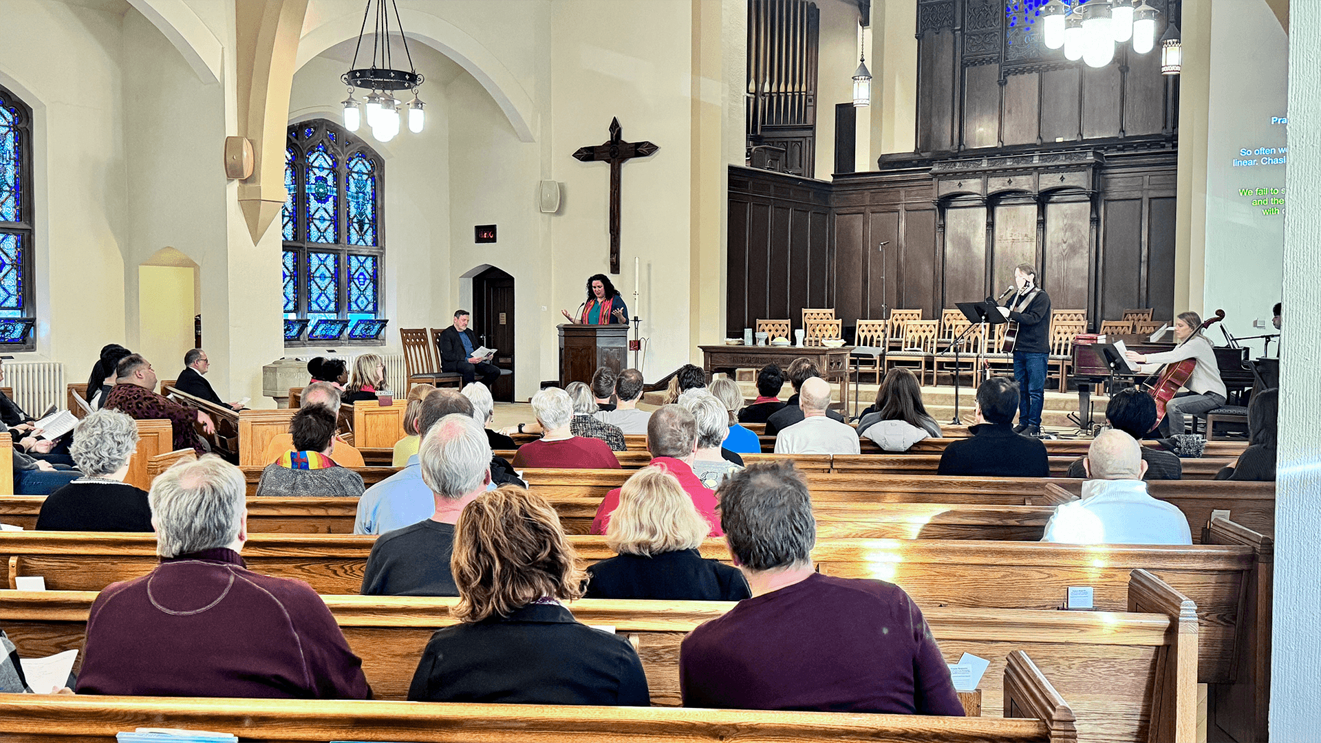 Speaker addressing a congregation at Grace Trinity Church from the pulpit, with a large wooden cross and stained glass windows in the background.