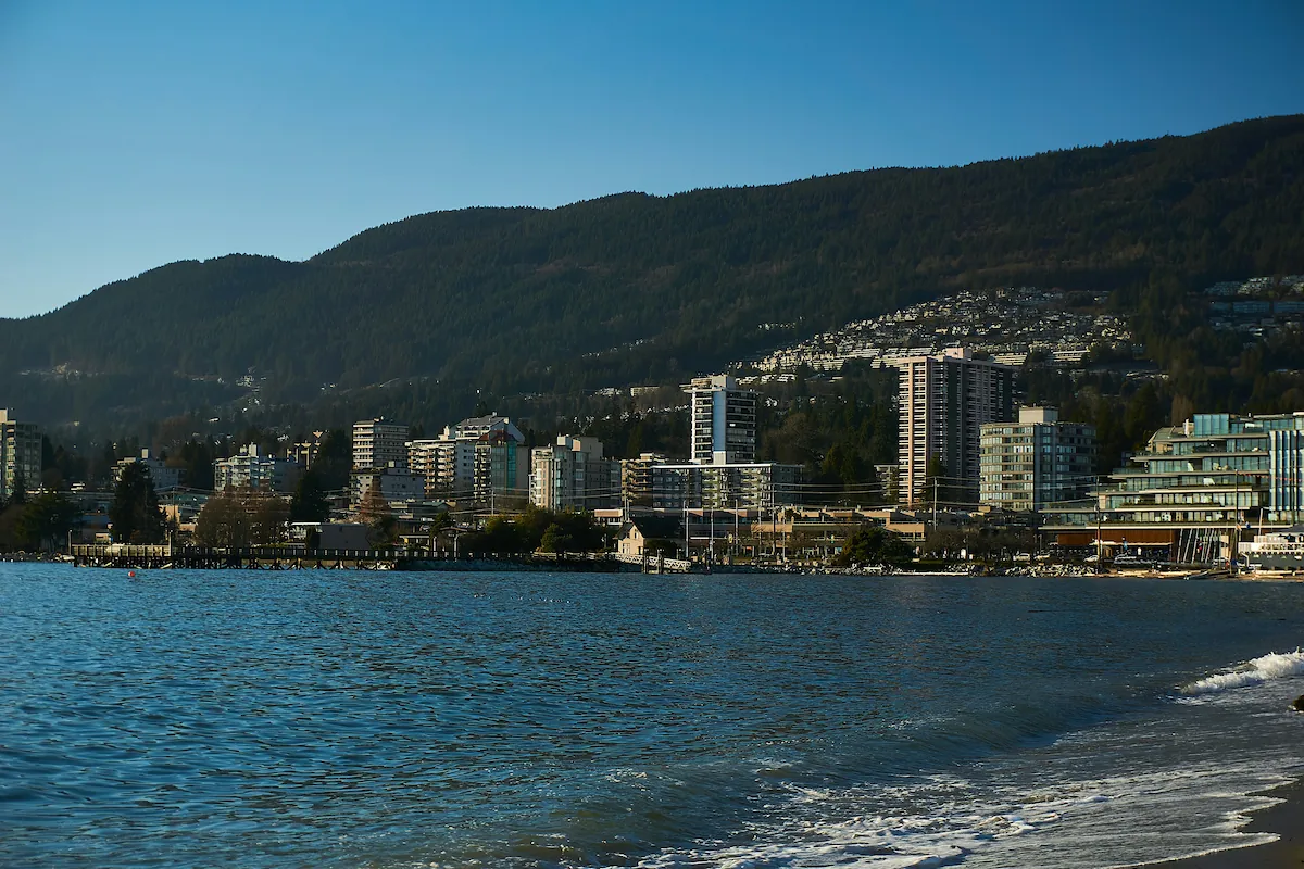 Waterfront view of North Vancouver with residential high-rises, forested mountains, and shoreline under clear blue skies.