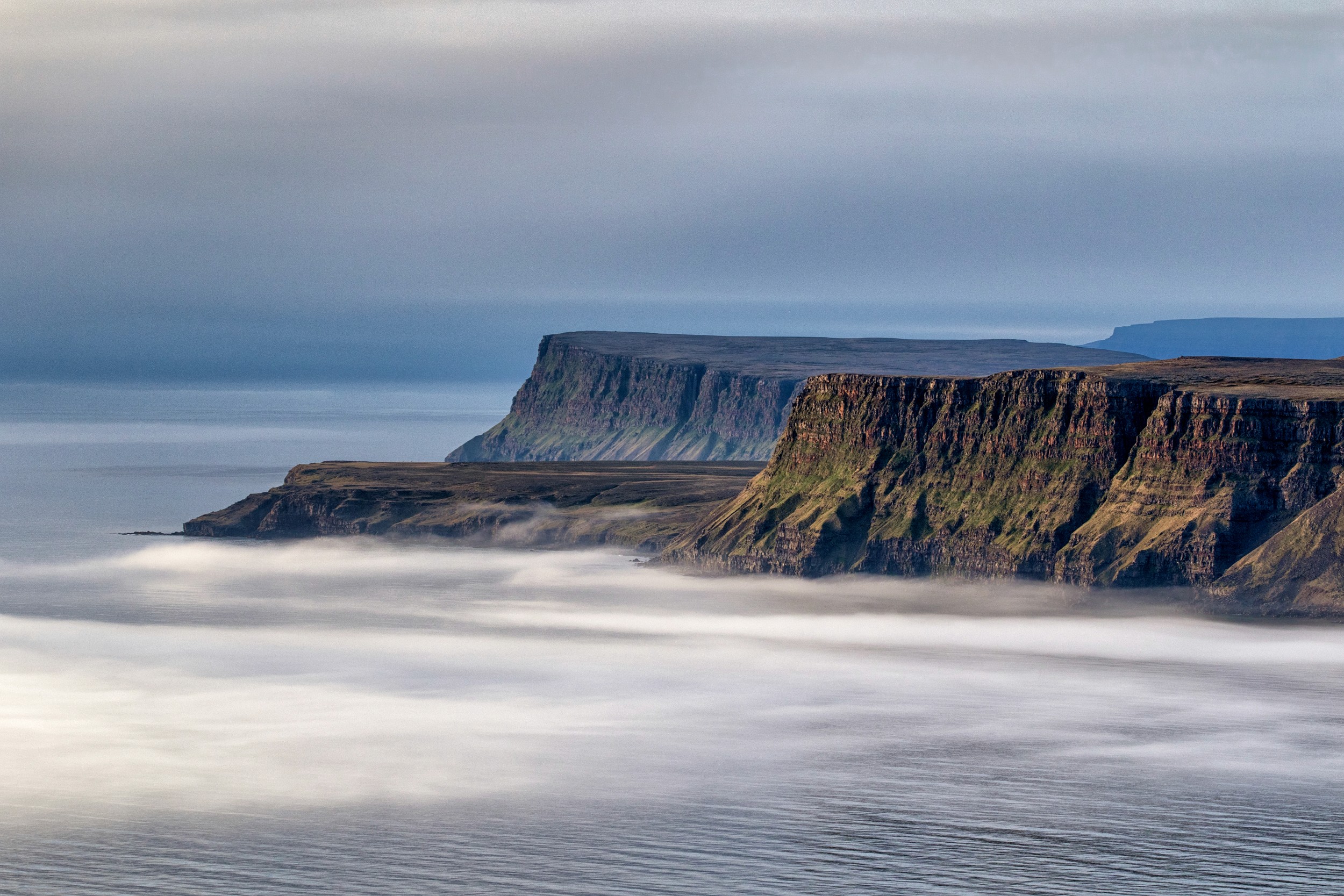 Dramatic cliffs rise above a misty shoreline, softened by long exposure under a moody sky