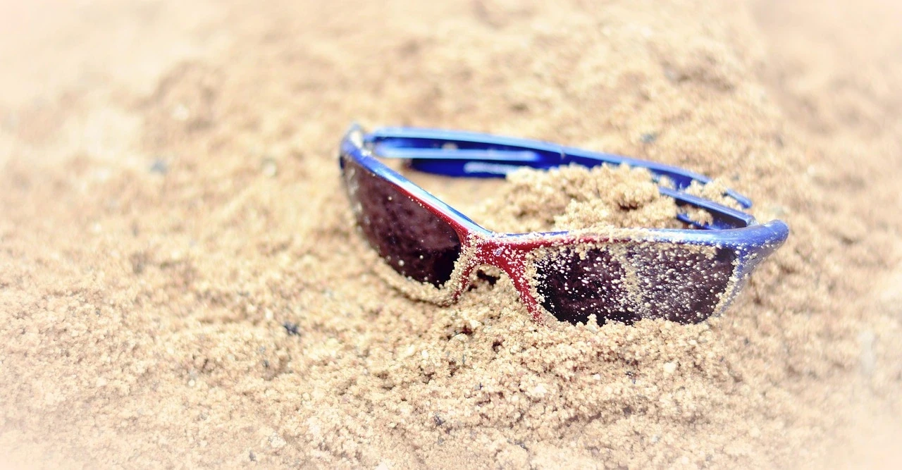 Gafas de sol de color azul y rojo cubiertas de arena en una playa, representando la tranquilidad de tener resueltas las opciones de pago en Brasil durante las vacaciones