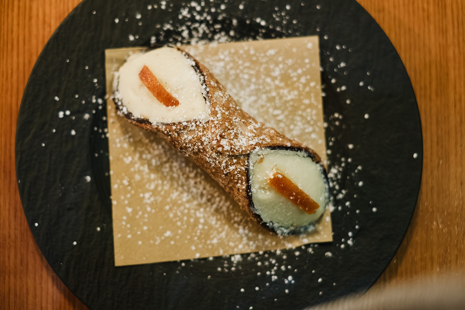 A single cannoli dusted with icing sugar, filled with ricotta cream and topped with small pieces of candied fruit, served on a black plate with parchment beneath it.