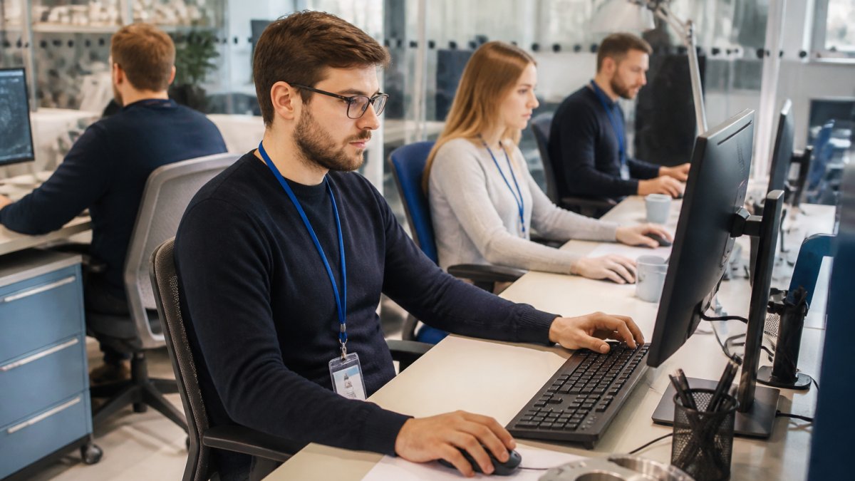 Engineering team members working at workstations in a secure office environment, reviewing CAD designs on multiple monitors with security badges visible