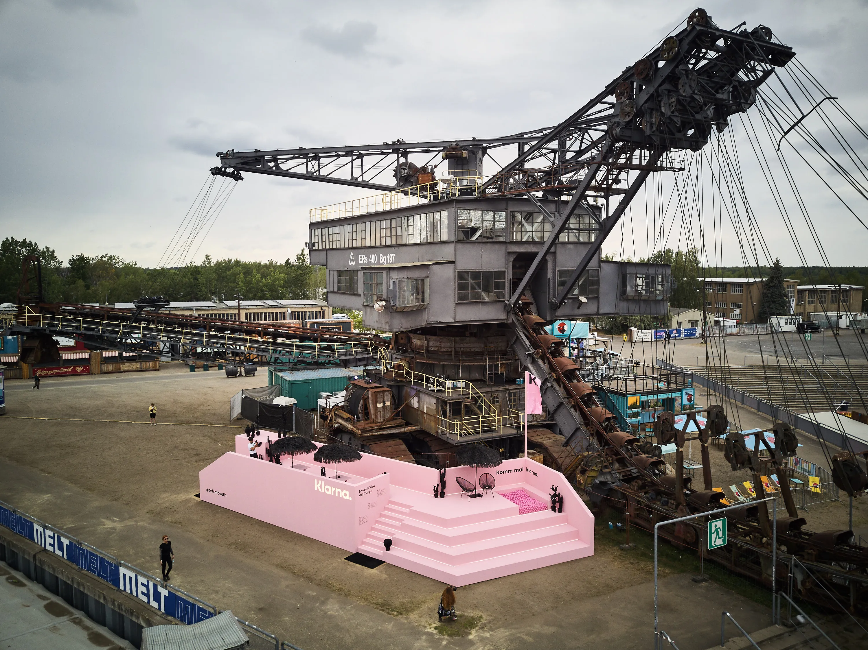 Aerial view of the pink Klarna platform with grandstand steps in front of a giant rusty bucket-wheel excavator at Melt Festival.