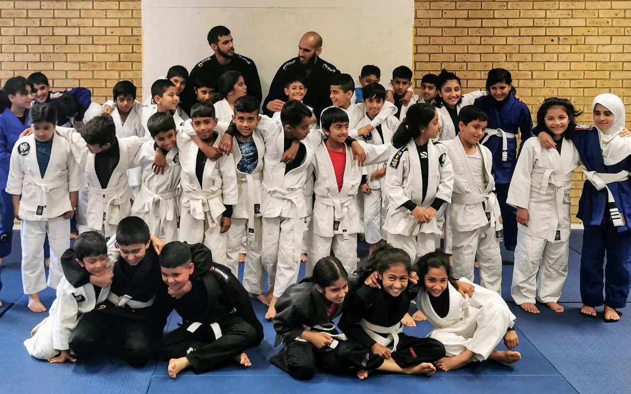Children and teenagers in white, blue and black judo and jiu-jitsu suits gathered together on blue mats, with two instructors standing behind them.