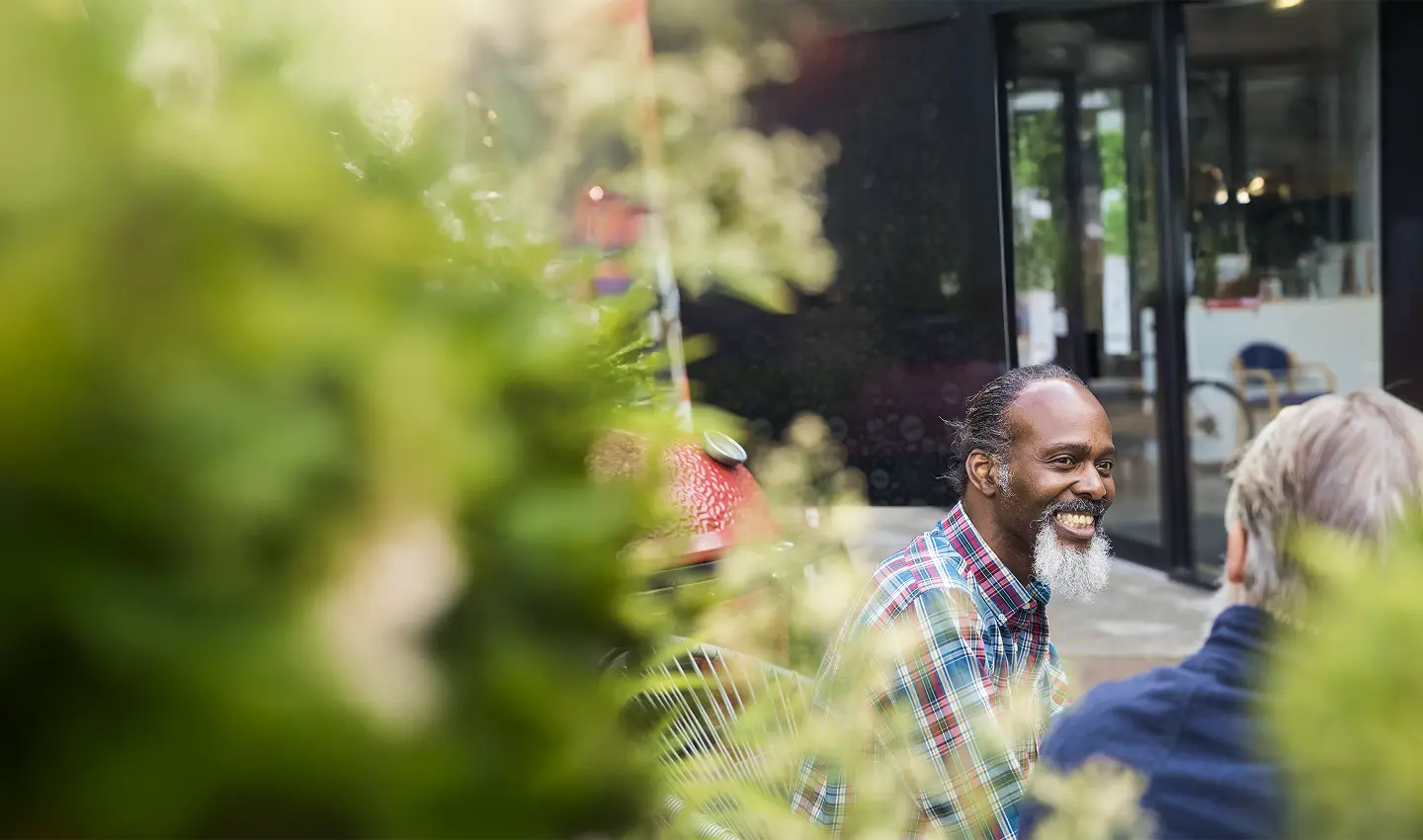 A group of people smiling and interacting outdoors, partially obscured by greenery in a vibrant setting.