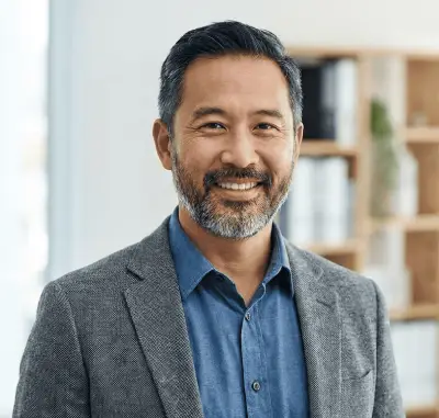 A smiling man in a blazer stands in an office setting with shelves in the background.