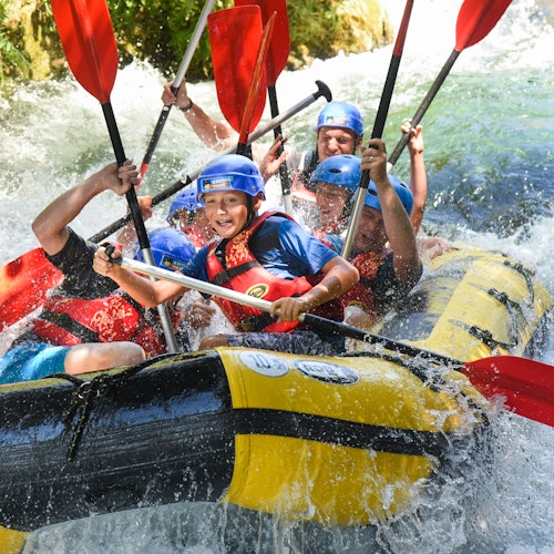 En grupp människor i hjälmar och flytvästar njuter av forsränning, paddlarna upphöjda, plaskar genom det vilda vattnet.