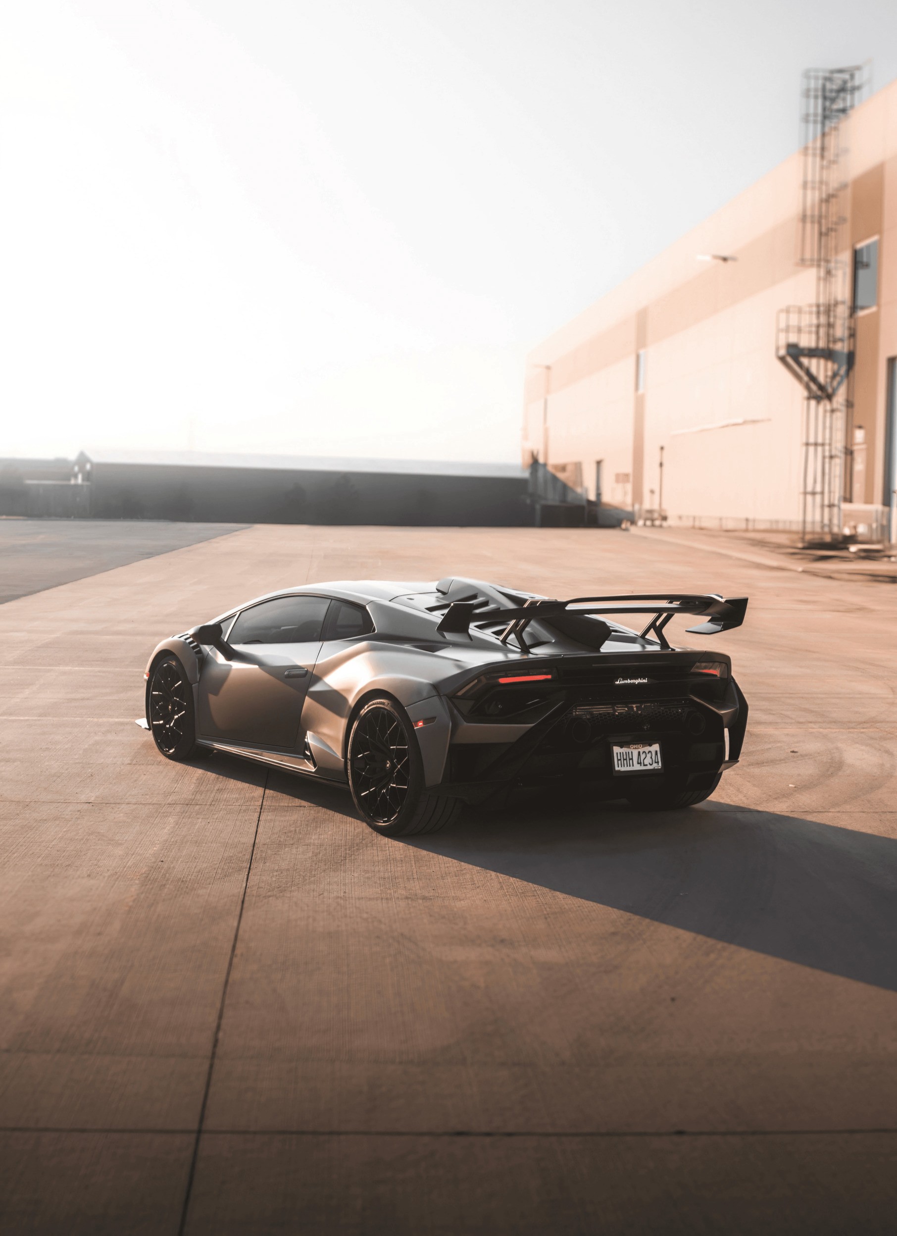 A sleek, modern sports car with a prominent rear spoiler parked on an expansive concrete lot beside a large industrial building under the soft glow of the afternoon sun.