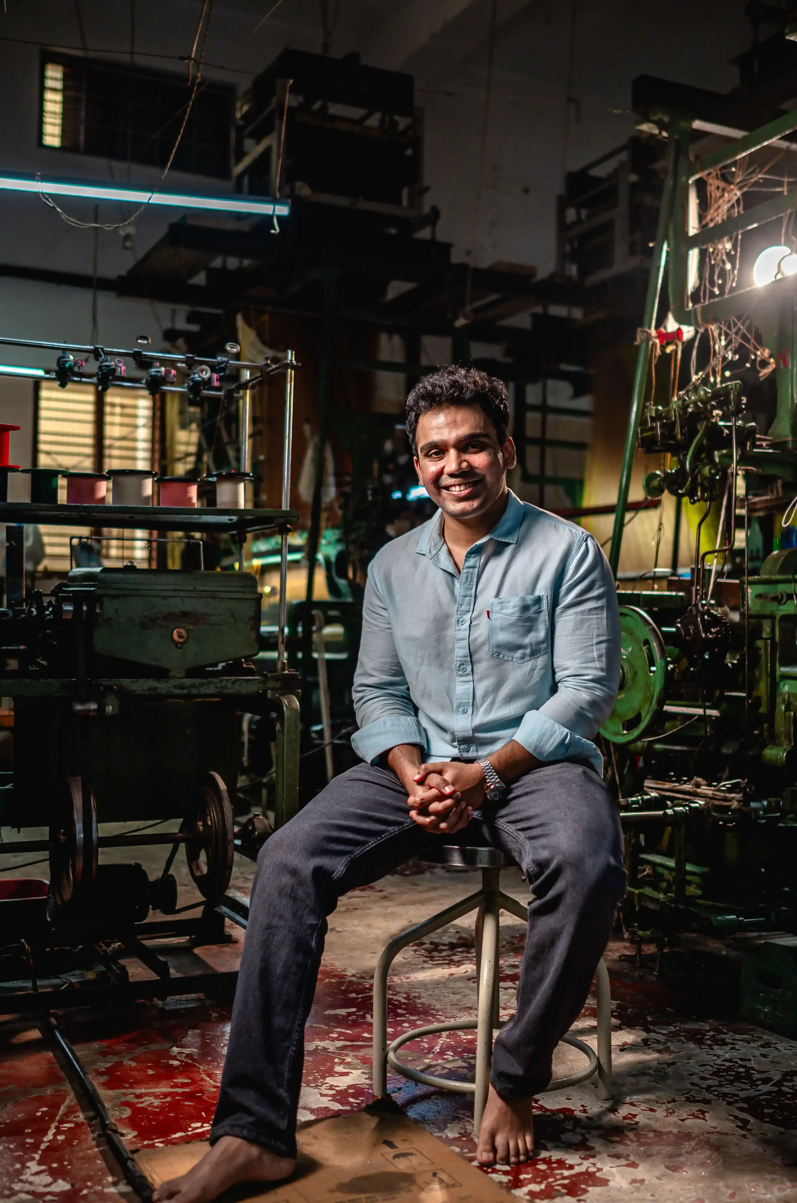 Shreyas Jeevan, founder of That Saree Shop, sitting on a chair inside the brand’s weaving factory, surrounded by handcrafted fabrics and artisans’ tools, reflecting his dedication to preserving traditional craftsmanship and quality.