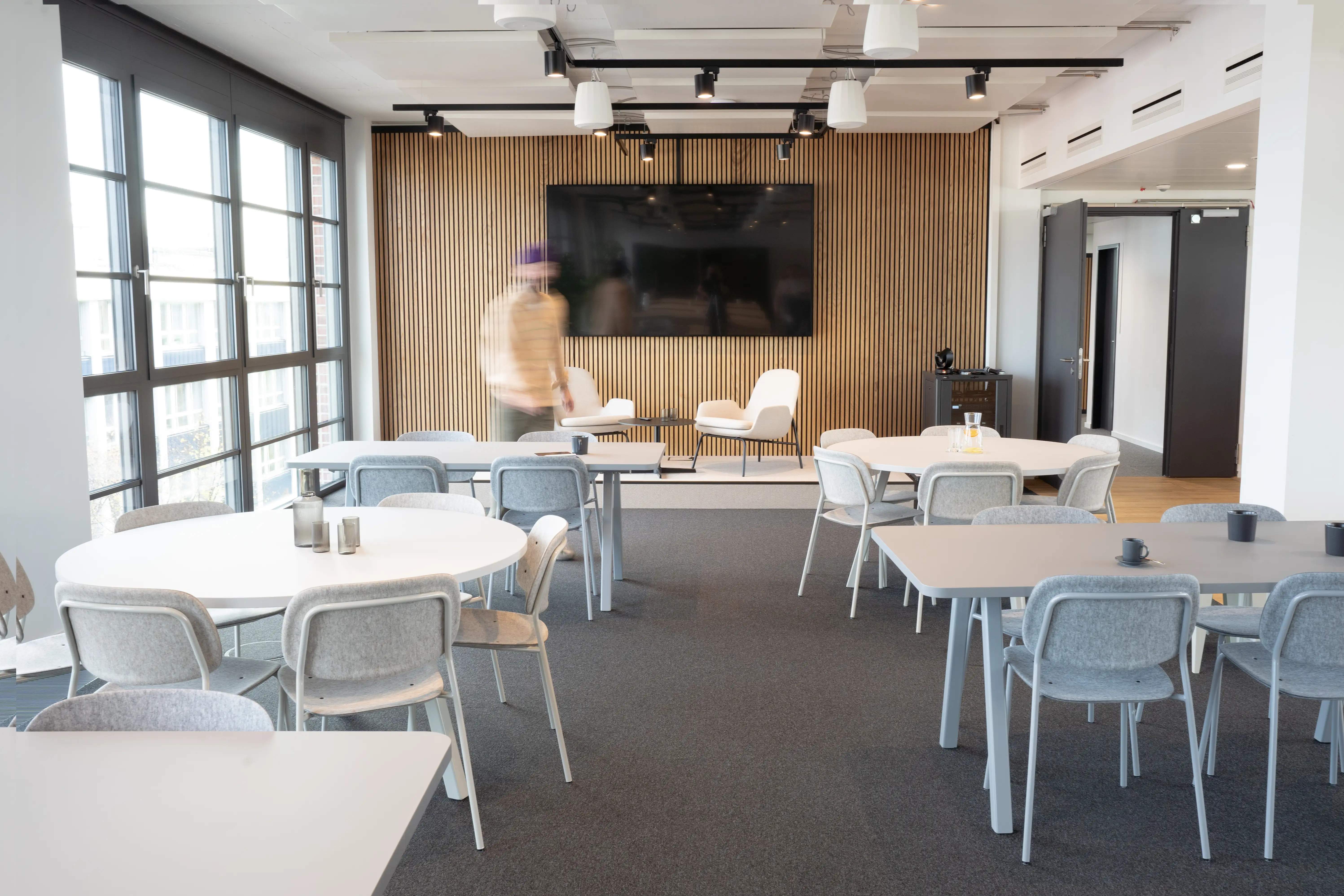 Work café at AI START-UP MUNICH with white round and rectangular tables, grey felt chairs, an oak slat media wall with large TV and white lounge chairs, and black track spotlights.