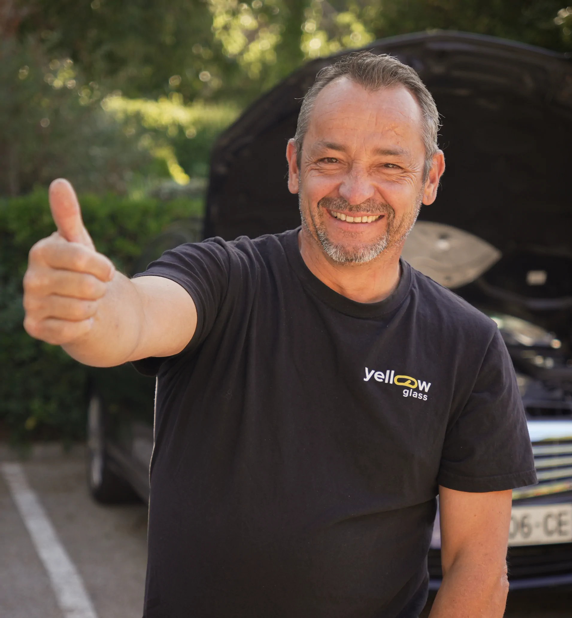 Un homme souriant donne son pouce en l'air, devant une voiture avec le capot ouvert. Il porte un t-shirt noir.