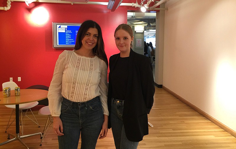 A photo of two women standing in an exhibition room, near a table with chairs.