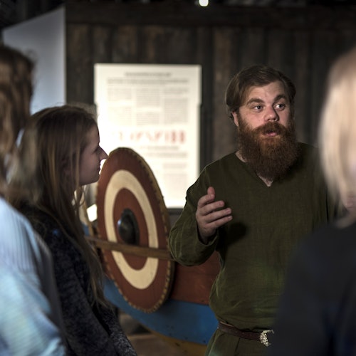 Egy szakállas férfi, zöld tunikában, gesztikulálva beszél egy kis csoport emberhez egy viking hajó kiállítás közelében.