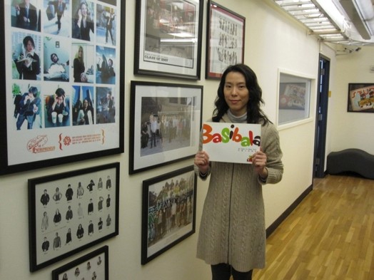 A photo of a women holding a banner with colorful letter text that says: Basibala, while standing near a wall filled with pictures in frames.
