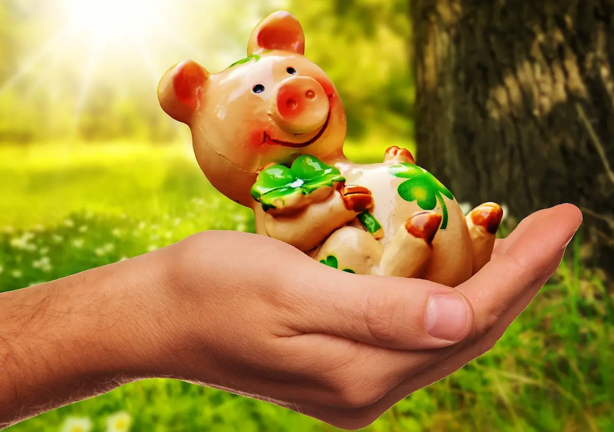 A smiling ceramic pig figurine holding a green four-leaf clover, resting in an open hand against a sunlit green meadow.