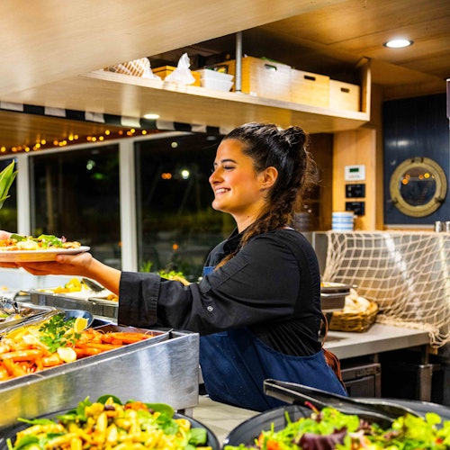 A smiling woman in a black uniform serves food at a salad bar in a restaurant with wooden decor and bright lighting.