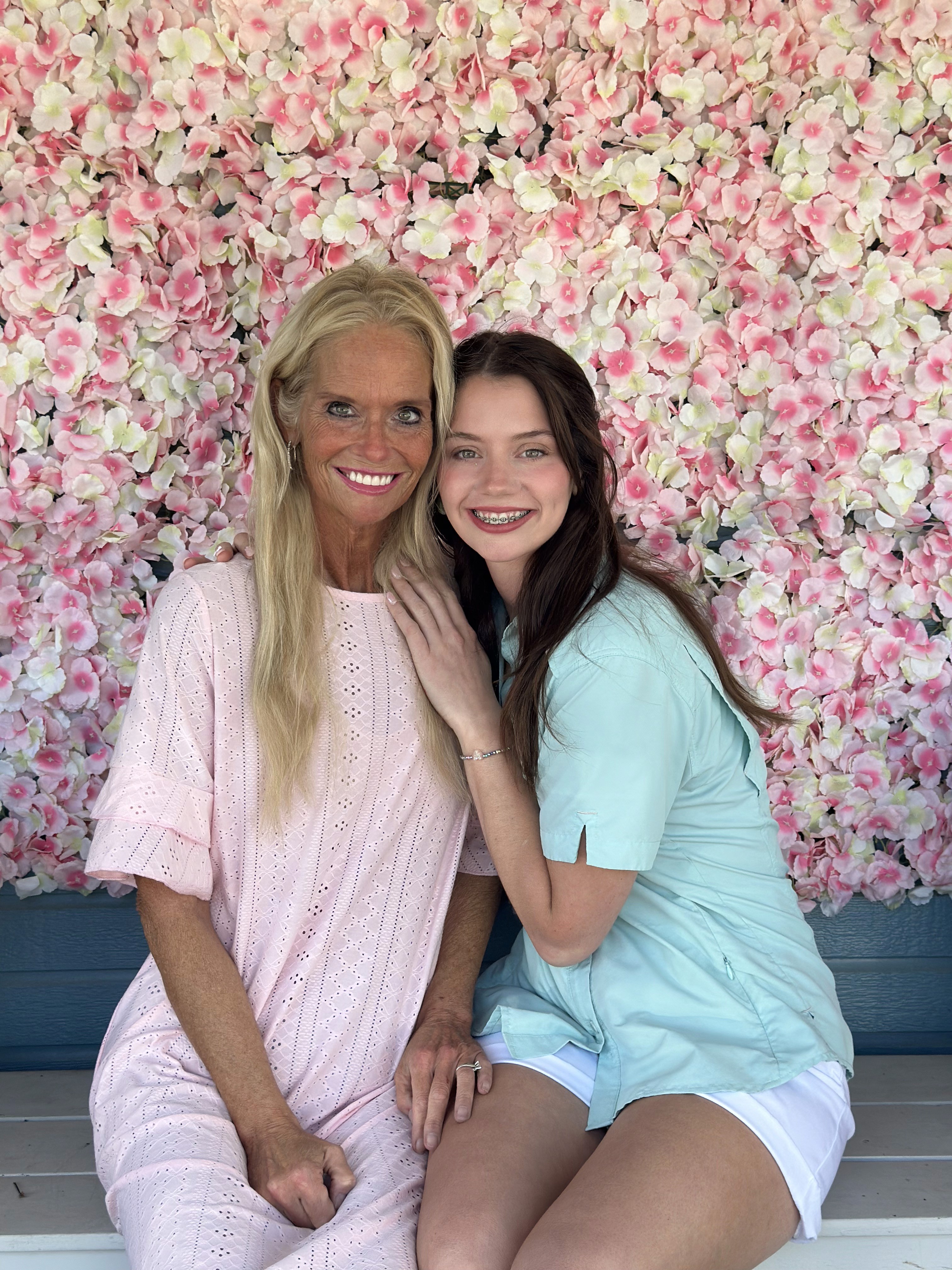 Two people are sitting in front of a vibrant pink and white flower wall, wearing light-colored summer outfits, smiling warmly at the camera.