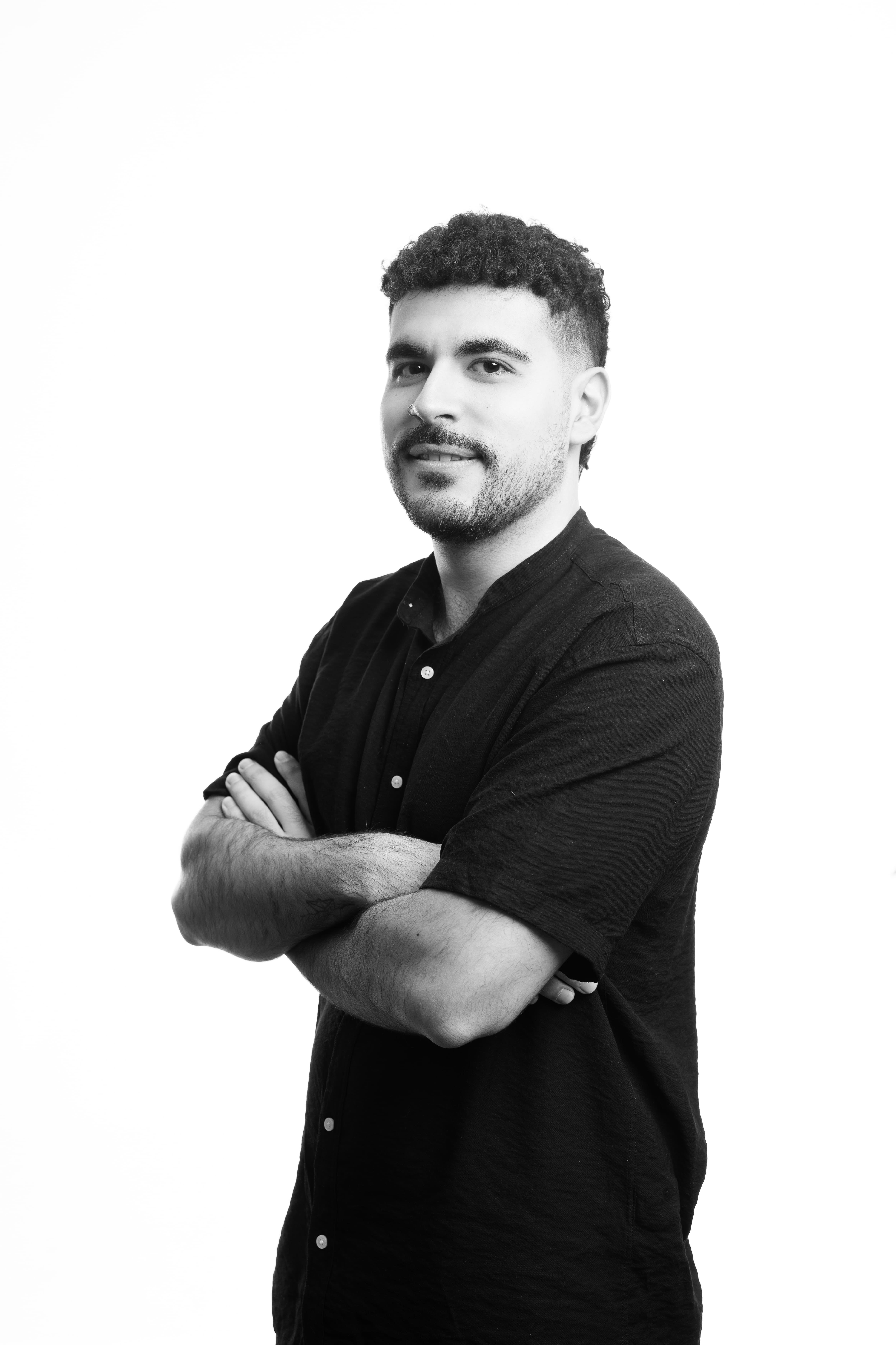A man with short curly hair stands confidently with crossed arms, wearing a dark button-up shirt. The background is plain white, creating a professional tone.