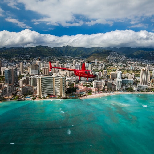 เฮลิคอปเตอร์สีแดงบินอยู่เหนือเมืองชายฝั่งที่มีตึกสูง ภูเขาสีเขียวอยู่เบื้องหลัง และน้ำทะเลสีฟ้าครามอยู่ด้านล่าง