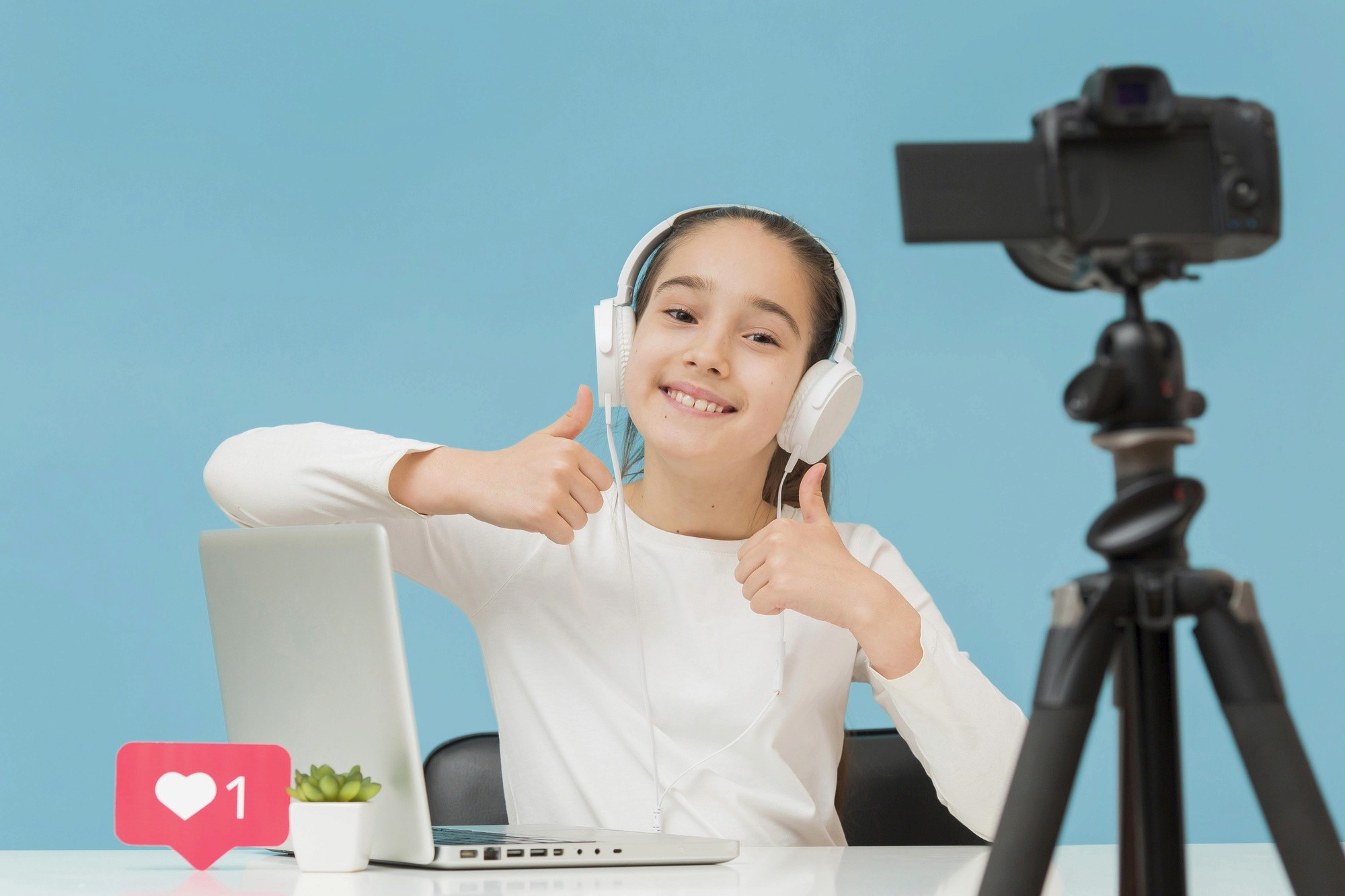 A girl wearing headphones and using a laptop, smiling and giving a thumbs up gesture.