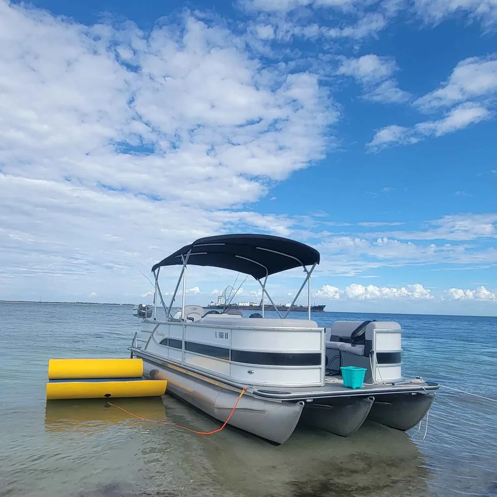 A pontoon boat is anchored near the shore in Bradenton, FL, with a clear blue sky, calm water, and a yellow floating device nearby.