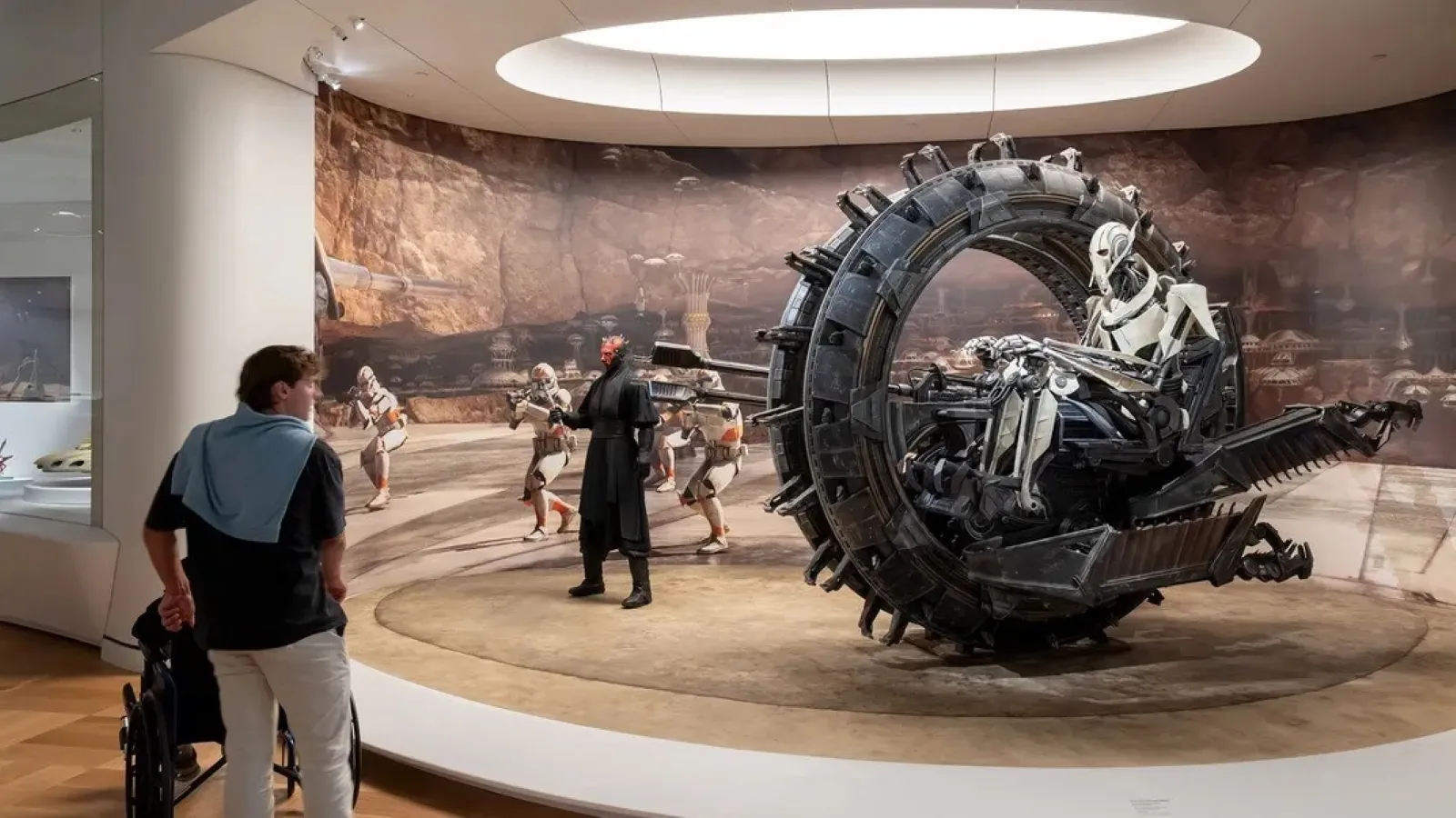 A man examines an intricate Star Wars exhibit featuring a large, circular vehicle with skeletal designs, surrounded by lifelike figures in dynamic poses, set against a desert backdrop inside a museum.