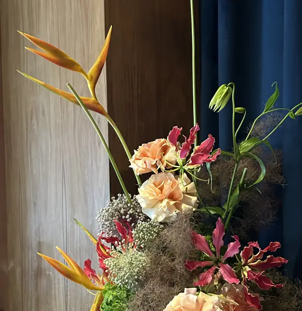 Arreglo floral contemporáneo con aves del paraíso, claveles durazno, gloriosas rojas y flores esféricas lilas sobre una repisa de madera clara frente a una cortina azul, junto a botellas de vidrio y vasos en una bandeja.