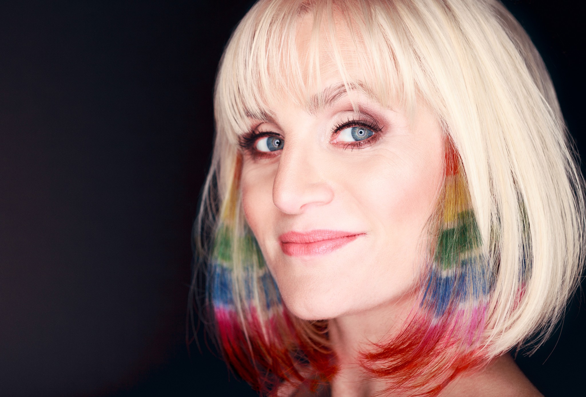 Beauty portrait of a blonde hair model with a sharp bob haircut and subtle rainbow color accents, photographed against a dark background.