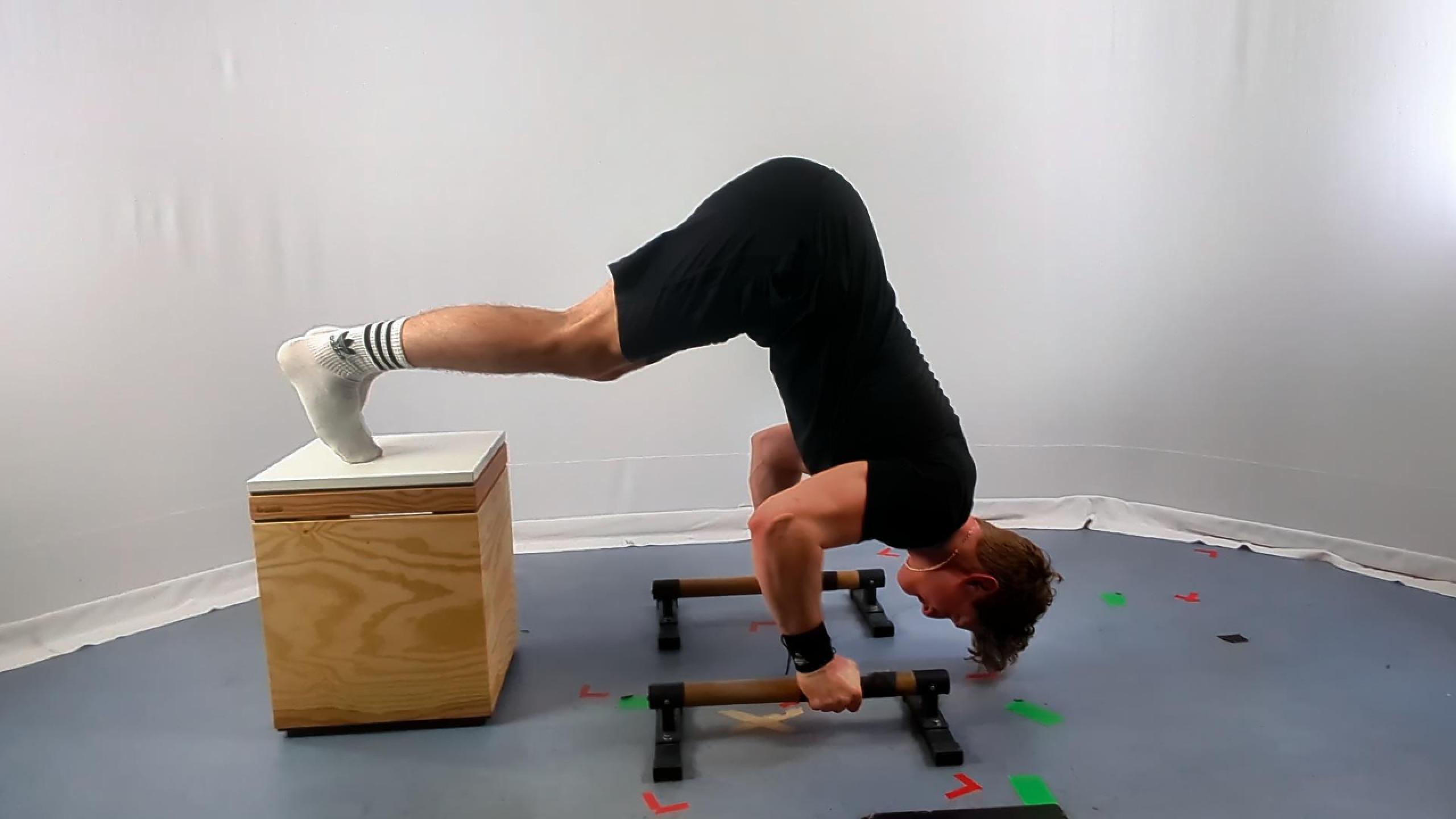 A man performs decline pike push-ups using wooden parallettes, balancing with his feet elevated on a box, demonstrating intermediate calisthenics strength and technique.