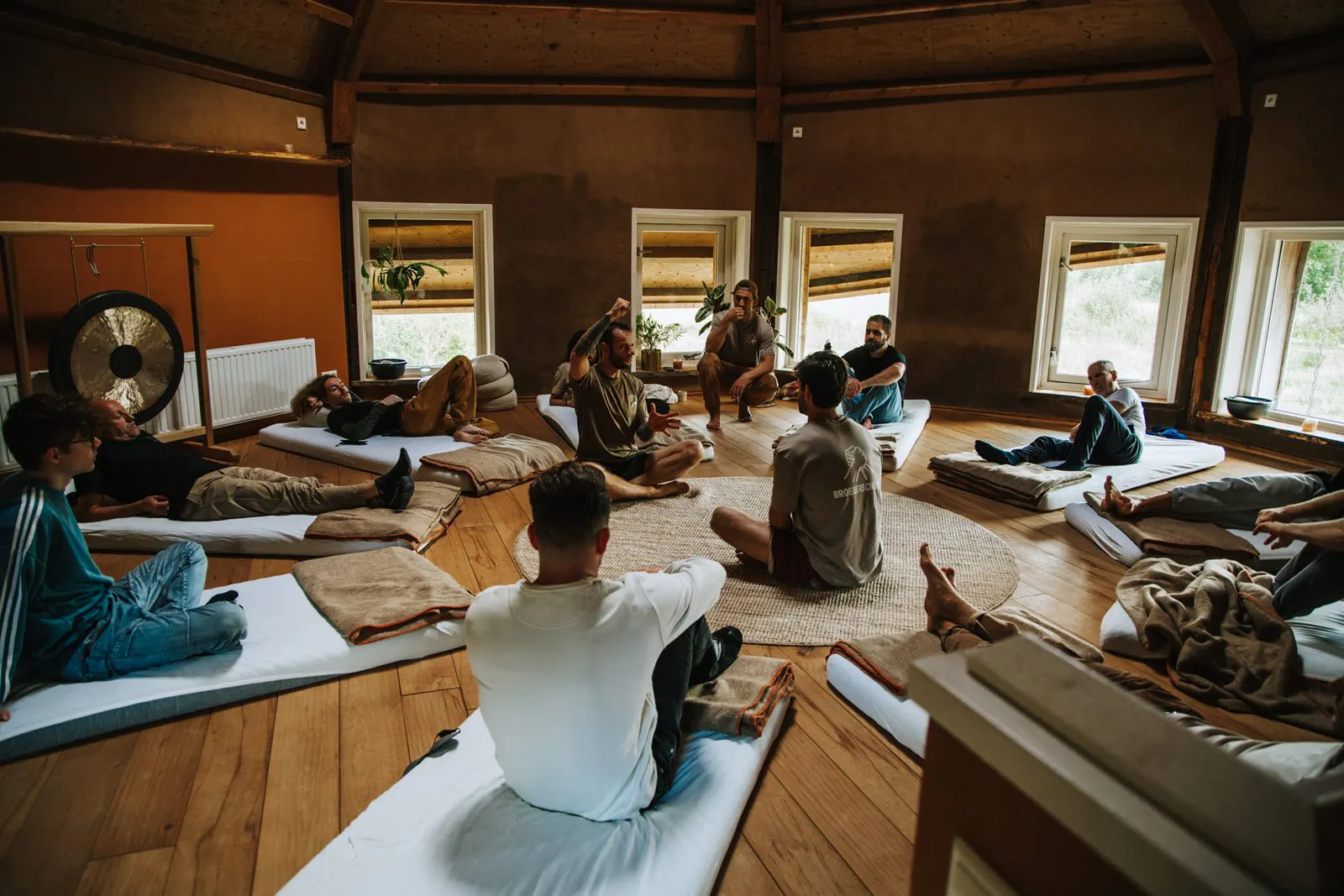Een groep mannen zit en ligt op witte matrassen in een kring om een rond kleed, met links een gong