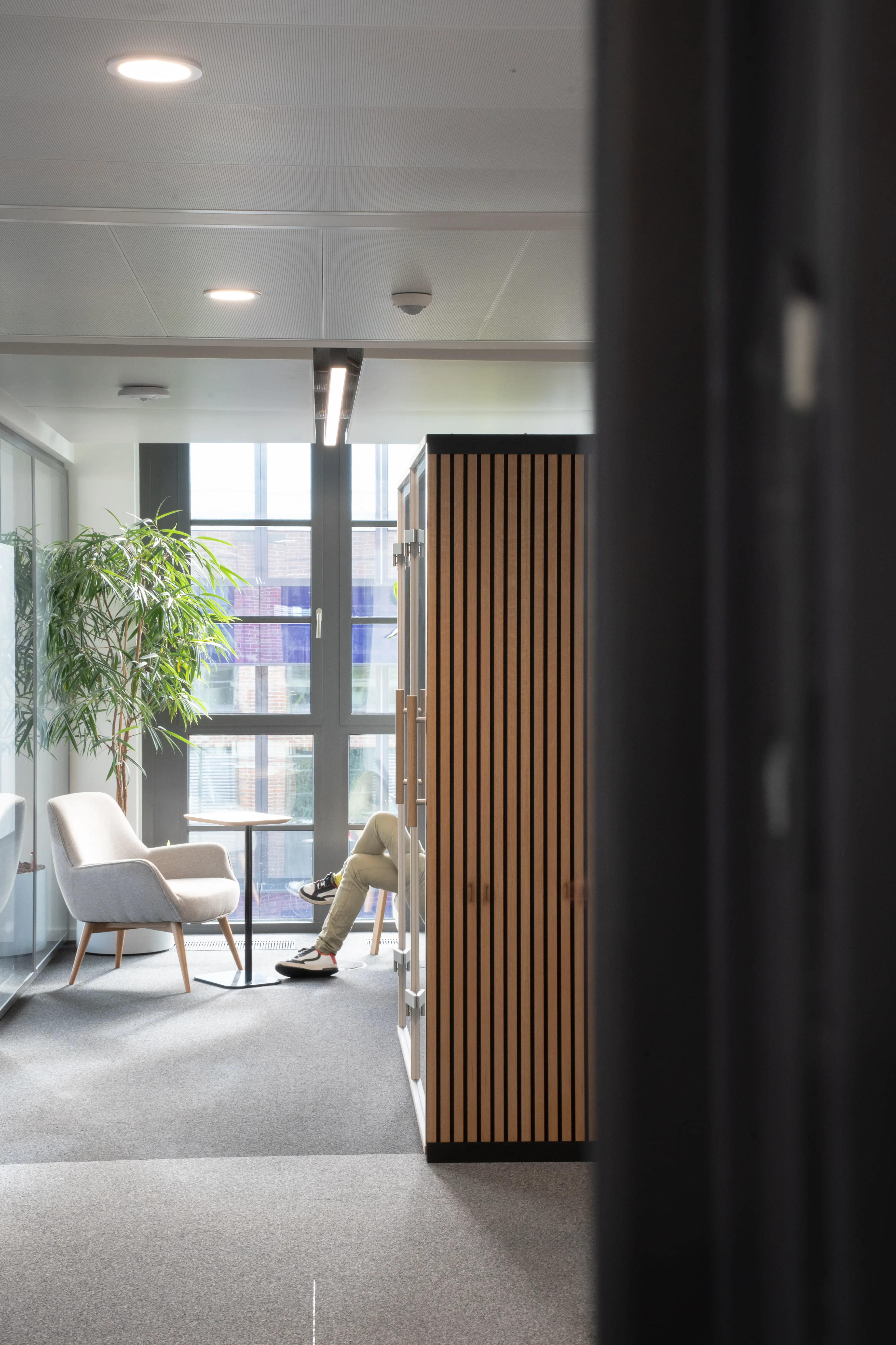View past an oak slat-clad cabinet at AI START-UP MUNICH into a daylight corner where a person sits in a cream armchair next to a small round table and an indoor tree.