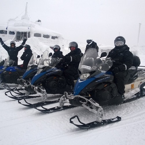 Lima orang mengenakan pakaian musim dingin duduk di atas snowmobile berwarna biru dan putih di lanskap bersalju, dengan latar belakang bangunan yang tertutup salju.
