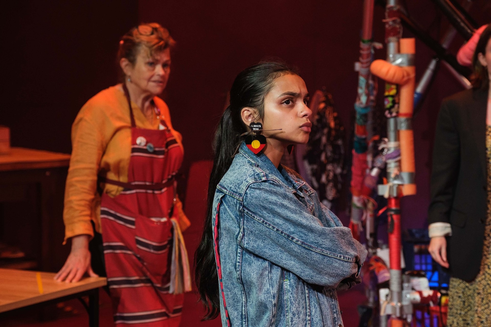 A performer in a denim jacket with bold earrings stands with arms crossed, looking determined. Behind them, another performer in an apron watches, framed by a cluttered workshop-like set.