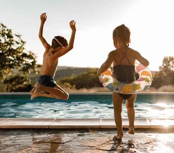 Two kids standing by a pool, and one of them jumping in