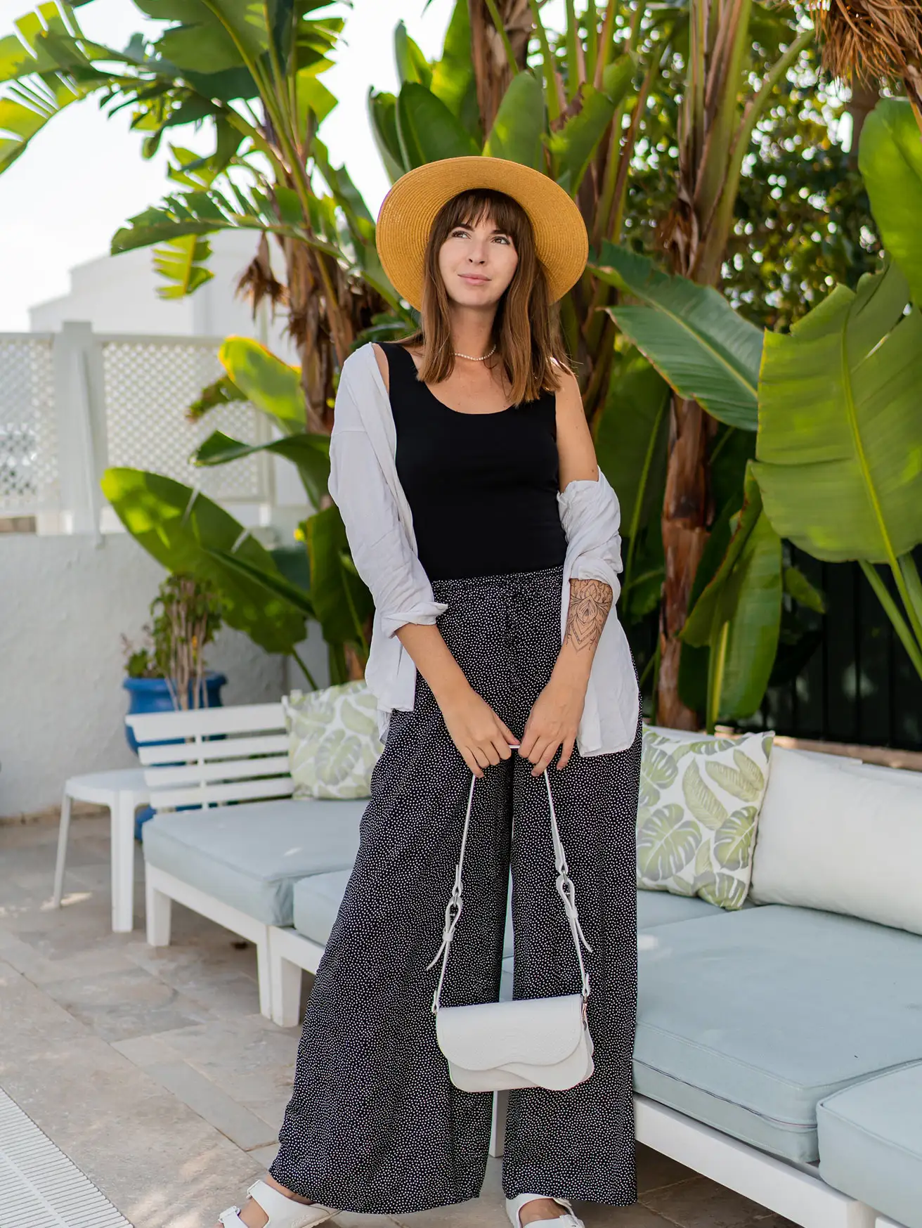 Woman in a black tank, polka-dot wide-leg trousers and straw hat on a sunlit terrace with palms