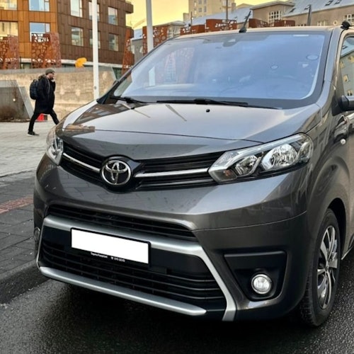 A gray Toyota minivan parked on a city street with buildings and a pedestrian in the background.