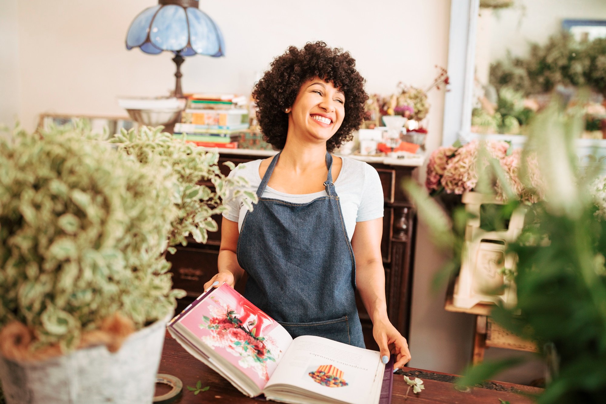 A smiling woman in a denim apron holding an open gardening or floral book, surrounded by plants and flowers in a flower shop or florist