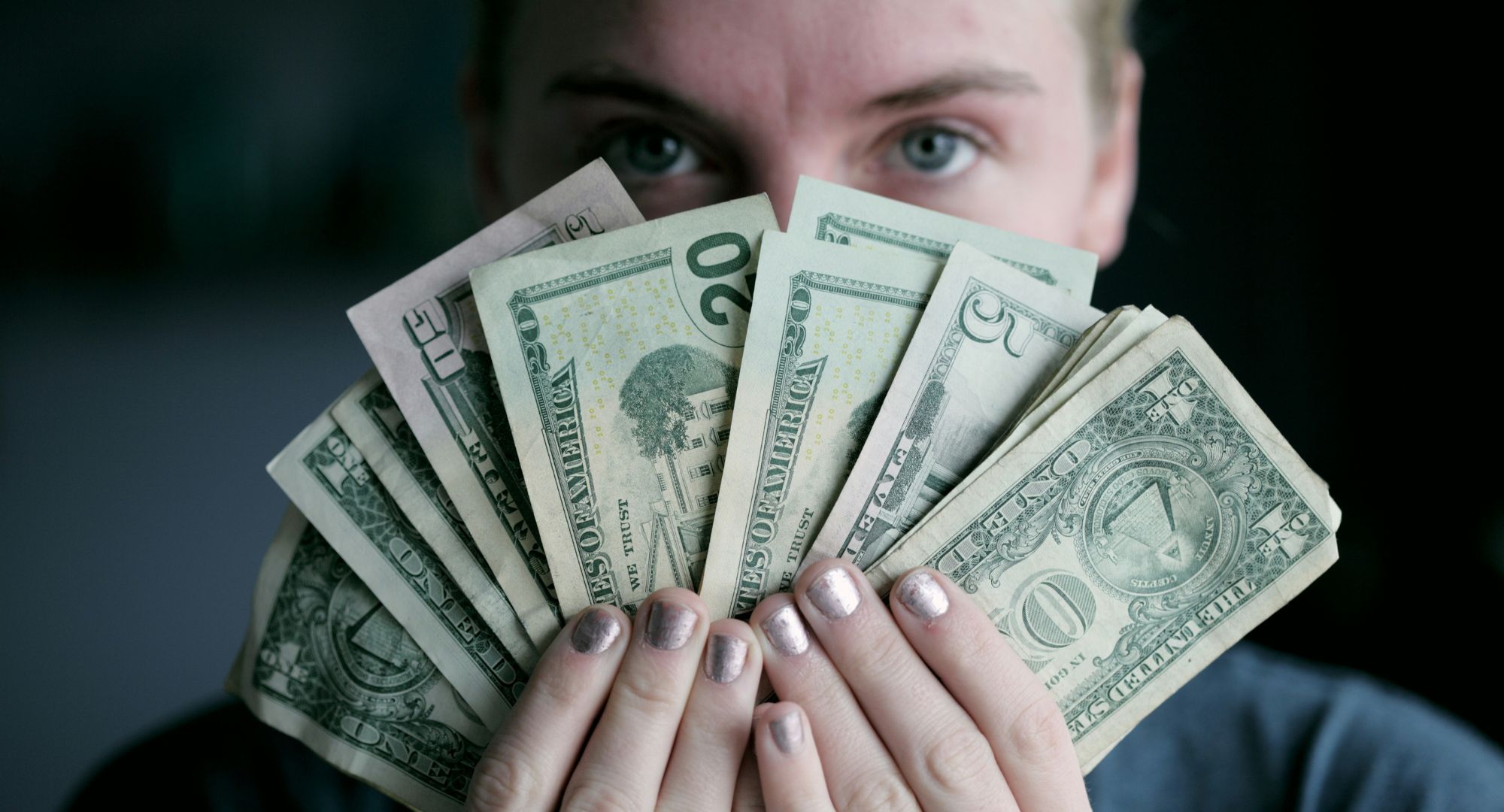 foto de pessoa segurando um leque de notas de dólares americano, o leque cobre quase todo rosto dela menos os olhos, ela olha para a câmera