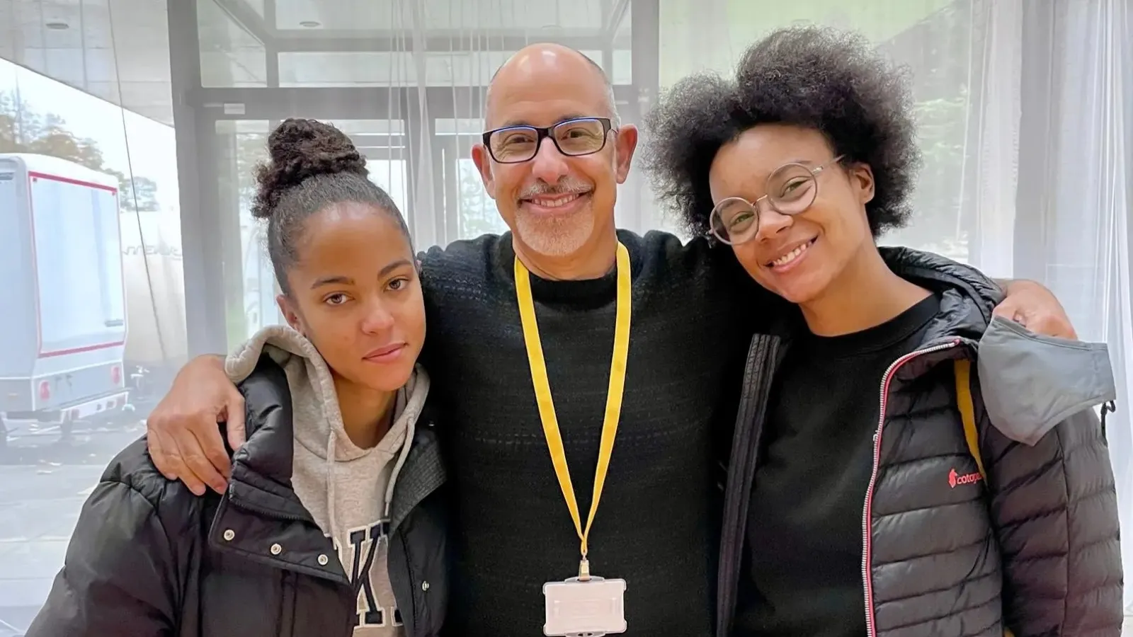 A man and two young women stood closely together and smiling in an indoor setting with large windows, one wearing a badge and two wearing winter jackets.