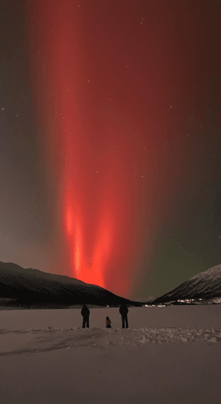 A deep red aurora rises behind a silhouette of mountains and people in a snowy field