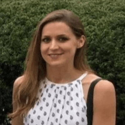Portrait of Kathryn Dean smiling outdoors, wearing a sleeveless patterned top with greenery in the background.